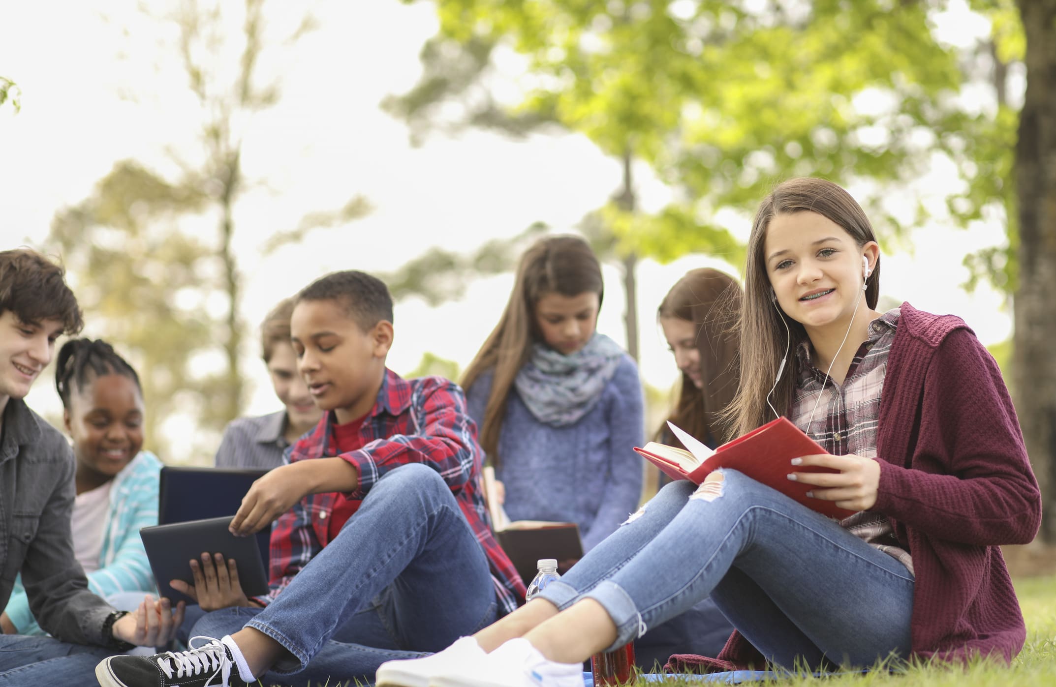 Multi-ethnic group of teenagers at park with friends. Pre-teenage and teenage group of boys and girls studying, hanging out together in local park or school campus with friends. Teen girl reading in foreground.